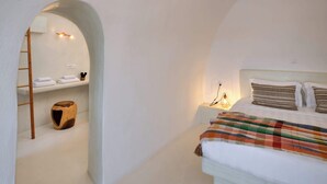 A bedroom with a bed, a nightstand, a lamp, a wooden stool, and a shelf with towels. - Amphora Meltemi Seaview Gem w/ Private Jacuzzi (Akrotiri)