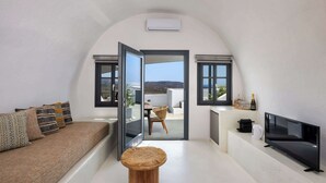 A modern living room with a sofa, a round wooden table, a flat-screen TV, and a view of the outdoors through a glass door. - Amphora Meltemi Seaview Gem w/ Private Jacuzzi (Akrotiri)