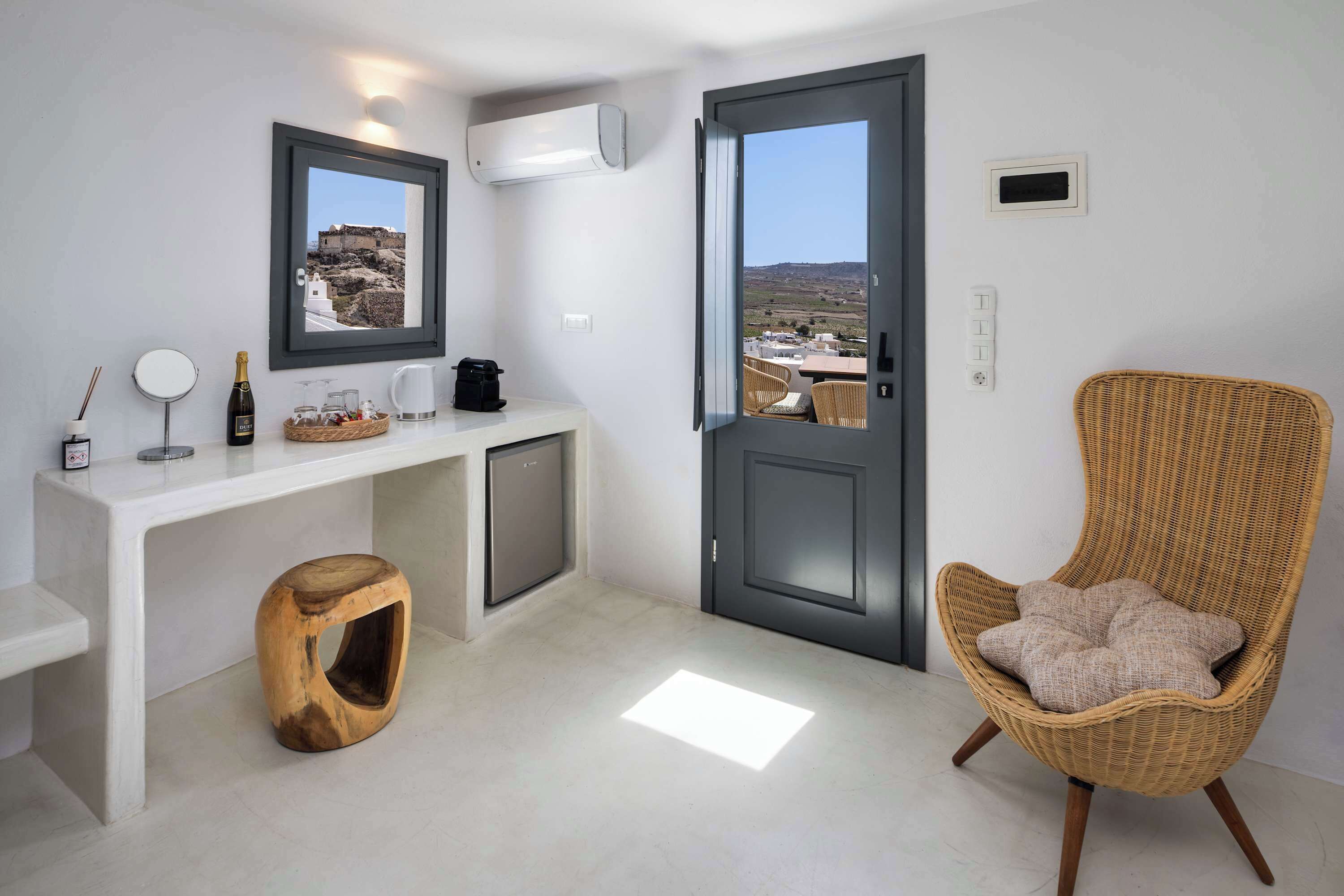 Habitació moderna i minimalista amb una taula consola blanca, una cadira de vimet i una porta que dona a un balcó amb vistes al paisatge.