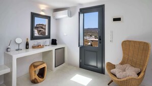 A modern, minimalist room with a white console table, a wicker chair, and a door leading to a balcony with a view of the landscape. - Amphora Thalassa Seaview Gem w/ Private Jacuzzi (Akrotiri)