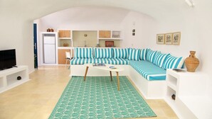 A living room with a sofa, a coffee table, a TV, and a kitchen area in the background. - Orasis House – Aegean Cave Retreat with Pool (Akrotiri)