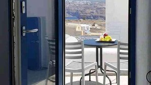 An open door leading to a balcony with a table and chairs, a view of the landscape, and a blue door frame. - Orasis House – Santorini Retreat w/ Pool & Views (Akrotiri)