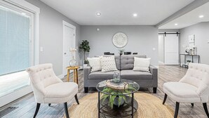 Una sala de estar moderna con un sofá gris, dos sillones, una mesa de centro de vidrio y un ventanal con persianas.