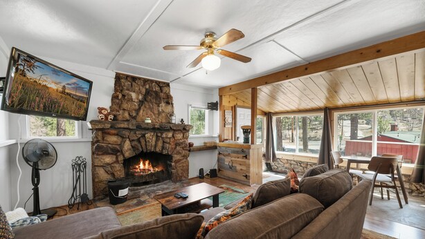 A cozy living room with a stone fireplace, a ceiling fan, and a large window overlooking the outdoors. - StoneHauz Cabin built in 1926 (Big Bear Lake)