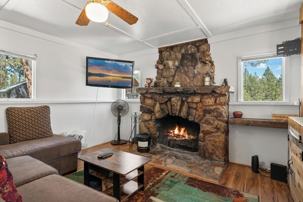 A living room with a stone fireplace, a television mounted on the wall, and a large window offering a view of trees. - StoneHauz Cabin built in 1926 (Big Bear Lake)