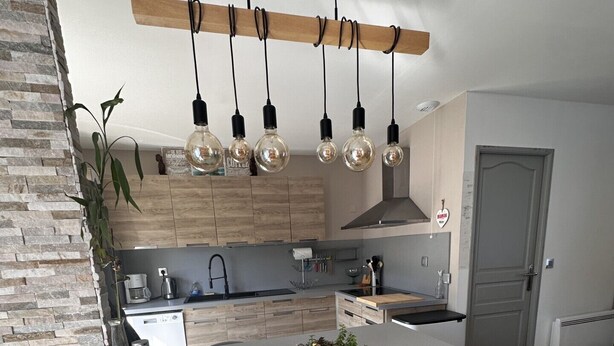 A modern kitchen with a large island, black bar stools, hanging light bulbs, and a stone wall.