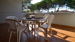 Une terrasse avec une table ronde et six chaises en plastique blanches, un saladier rempli de fruits et un vase contenant une seule fleur.