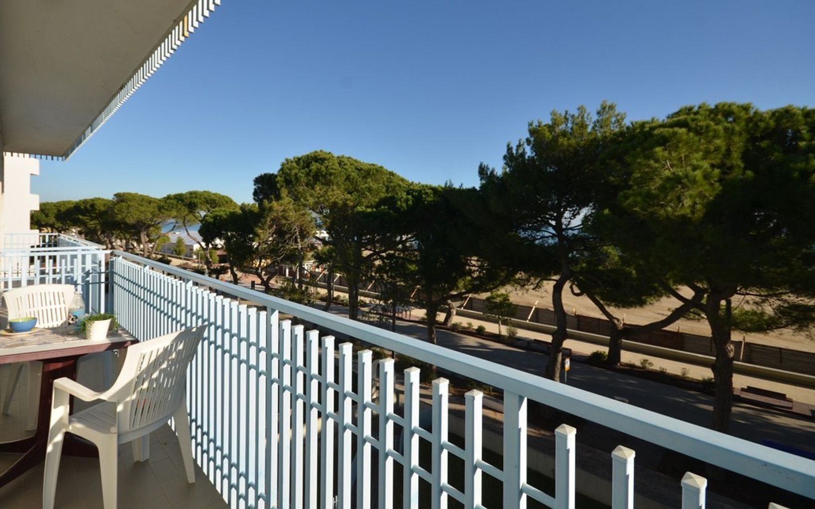 A balcony with a white railing overlooking a street lined with trees and a clear sky.