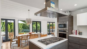 A modern kitchen with a central island, stainless steel appliances, and a view of the garden through large sliding doors.
