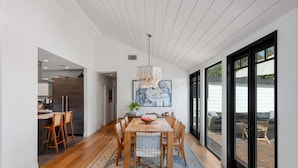 A modern dining area with a wooden table and chairs, a hanging light fixture, and a large window with a view of the outdoors.