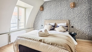 Un dormitorio con techo inclinado, una cama grande con cabecera de madera, papel tapiz estampado y una ventana que proporciona luz natural.
