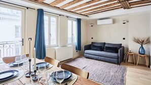 Comedor con mesa de madera dispuesta para cuatro, sofá gris y vista de la ciudad a través de amplios ventanales.