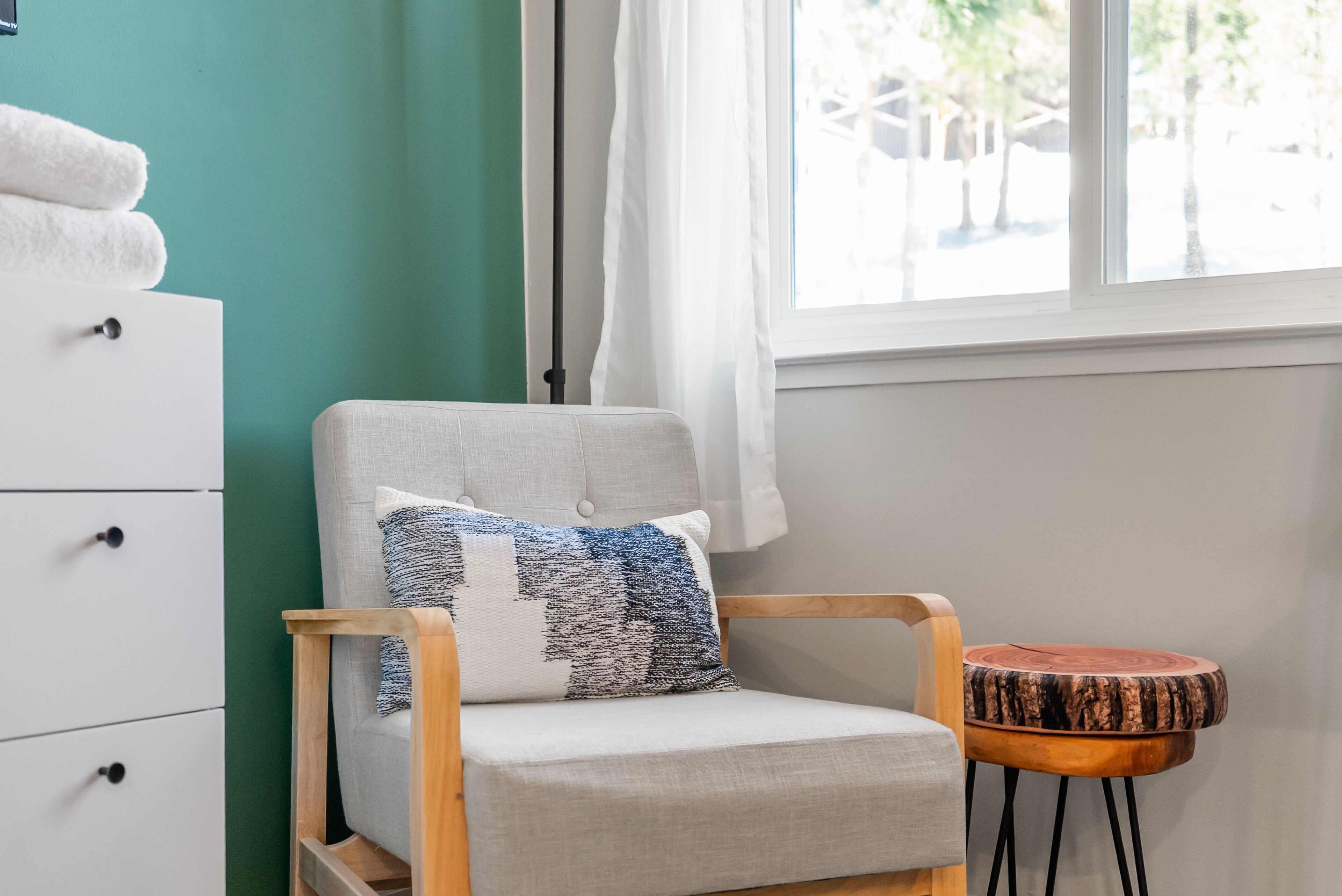 A room with a white chair, a small round wooden table, and a white dresser with drawers.