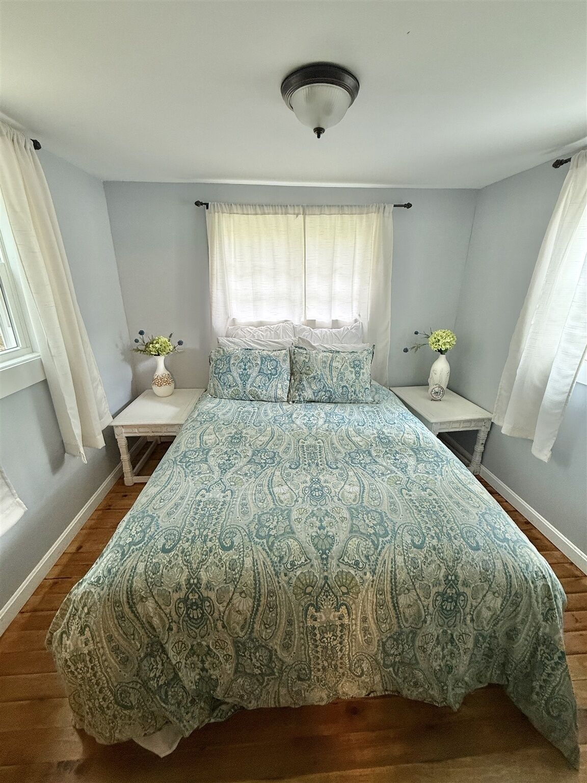 Un dormitorio con una cama, dos mesitas de noche, una ventana con cortinas y una lámpara en el techo.