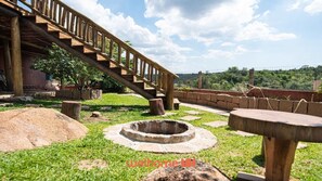 A wooden staircase leading up to a building, surrounded by stone walls and a grassy area with a fire pit.