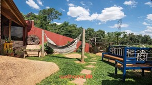 A rustic outdoor area with a hammock, a wooden bench, and a stone pathway leading to a building with a red wall and a satellite dish.