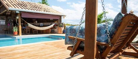 A wooden deck with a pool, a swing chair, and a building with a tiled roof.
