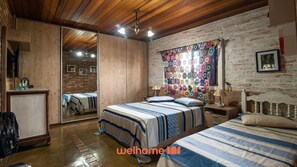 A bedroom with a wooden ceiling, a brick wall, a large mirror, a bed with striped bedding, a nightstand, and a colorful floral tapestry.