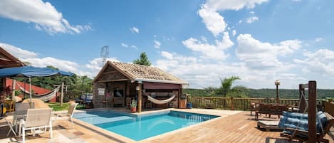 A wooden deck with a swimming pool, a small stone building with a tiled roof, and a hammock.