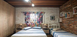 Un dormitorio con pared de ladrillo, una ventana con cortina de ganchillo colorida, dos camas con ropa de cama a rayas, mesitas de noche con lámparas y una estantería.