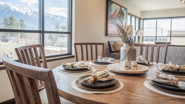 A dining table set with plates, napkins, and a vase with dried flowers, with a mountain view through large windows. - Downtown Penthouse Condo – For 6! (Columbia Falls)