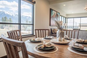 Una mesa de comedor dispuesta con platos, servilletas y un jarrón con flores secas, con vistas a la montaña a través de amplias ventanas.
