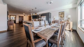 Ein moderner Essbereich mit einem Holztisch, gedeckt für sechs Personen, umgeben von Stühlen. Im Hintergrund ist eine Küche zu sehen, und auf der rechten Seite ein großes Fenster.