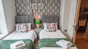 Ein Hotelzimmer mit zwei Betten, einer Wand mit geometrischem Muster, einem kleinen Tisch mit Lampe und Blick auf einen Korridor.