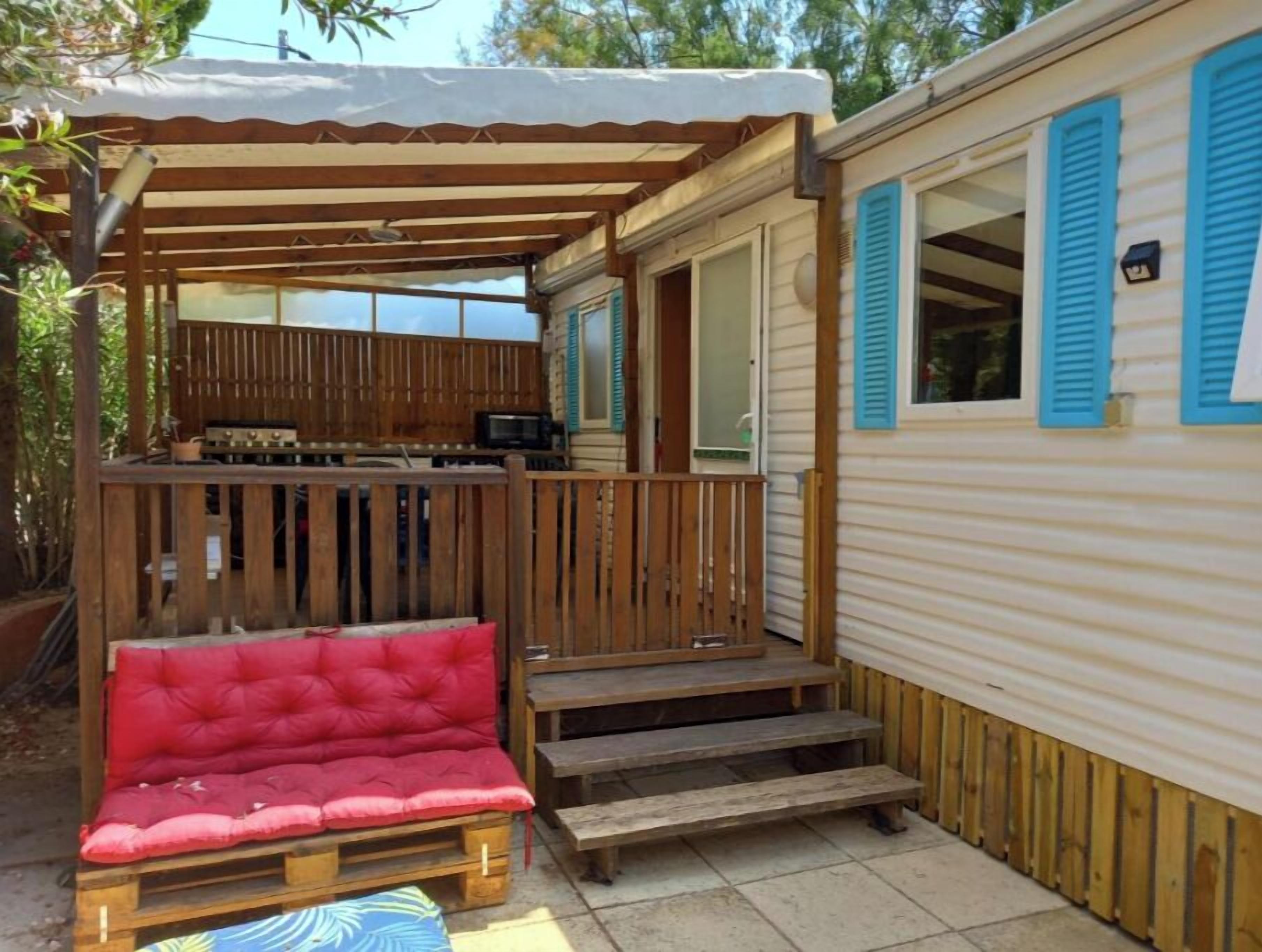 A covered outdoor seating area with a red cushioned bench and a wooden table, adjacent to a mobile home with blue shutters.