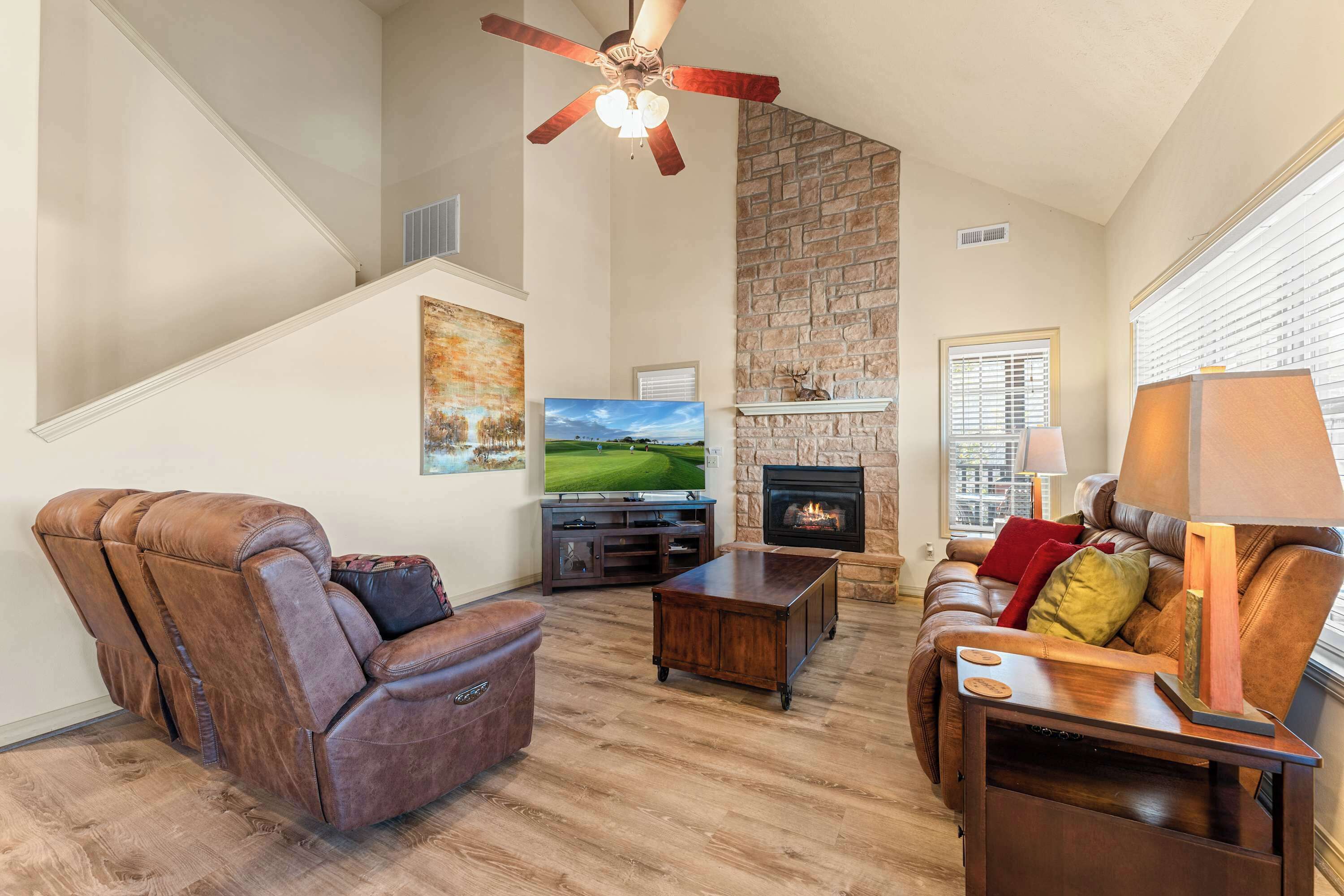 A spacious living room with a stone fireplace, leather recliner, wooden coffee table, and a ceiling fan.