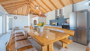 A spacious kitchen with a wooden ceiling, a long wooden table with cushioned benches, a stone fireplace, and stainless steel appliances.