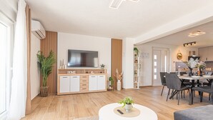 A modern living room with a flat-screen TV, a dining area with a table and chairs, and a kitchen in the background.