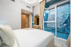 Une chambre d’hôtel moderne avec un grand lit, une petite table de chevet, un miroir et une vue sur le paysage urbain, visible à travers une porte vitrée.
