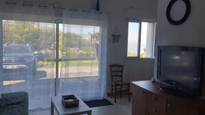 Een kamer met een televisie op een houten kast, een rieten mandje op een tafel, een stoel, en uitzicht op een geparkeerde auto door een raam met dunne gordijnen.