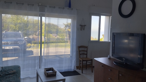 A room with a TV on a wooden cabinet, a wicker basket on a table, a chair, and a view of a parked car through a window with sheer curtains.