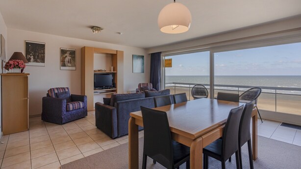 A modern living room with a dining area, a TV, and a large window with a view of the sea.