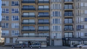 A multi-story residential building with balconies, a ground-level commercial space, and parked cars.