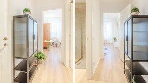 Un pasillo con piso de madera, una silla blanca, cerramiento de ducha de vidrio y un estante negro con una planta encima.