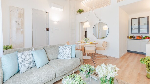 Una sala moderna con un sofá gris, una mesa de comedor con sillas, una zona de cocina con armarios y un espejo grande.