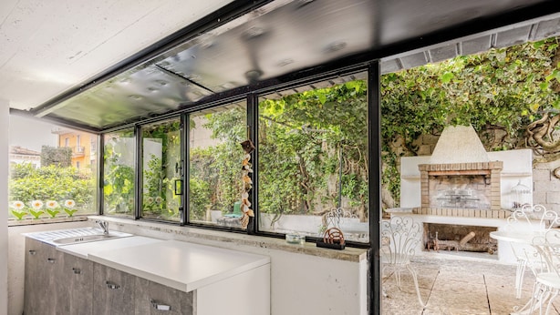 A modern kitchen with a large island, a built-in oven, and a view of a garden through large windows.