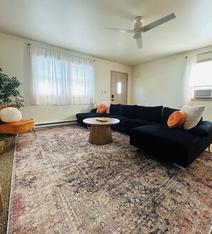 Ein Wohnzimmer mit einer schwarzen Couch, einem runden Couchtisch, einem Deckenventilator und einem gemusterten Teppich.
