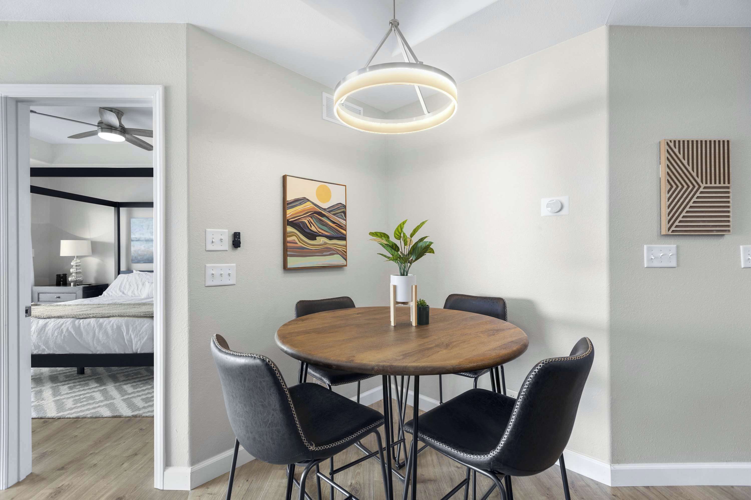 A modern dining area with a round wooden table, black upholstered chairs, a potted plant, and a contemporary pendant light.