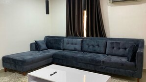 A dark blue sectional sofa with a remote control on a white coffee table in a living room with a large window and dark curtains.