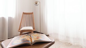 A wooden chair with a cushion, an open book with colorful illustrations, and a floor lamp in a room with a white wall and sheer curtains. - Italianway Easy - Palazzi 6 C (Milan)