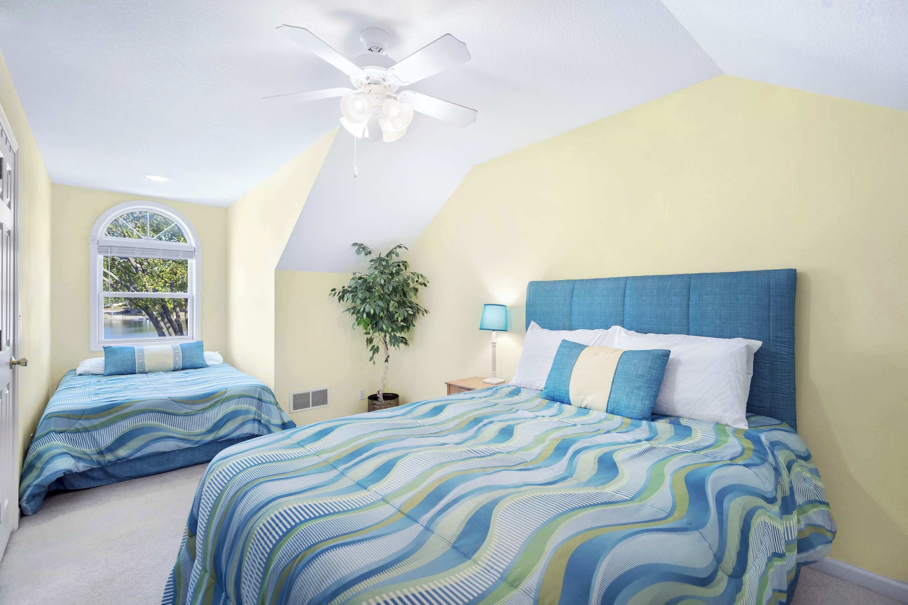A bedroom with two beds, a ceiling fan, a window, a plant, and a nightstand with a lamp.