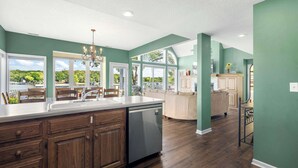 A spacious kitchen with a large island, wooden cabinets, and a dishwasher. There are large windows offering a view of the outdoors.