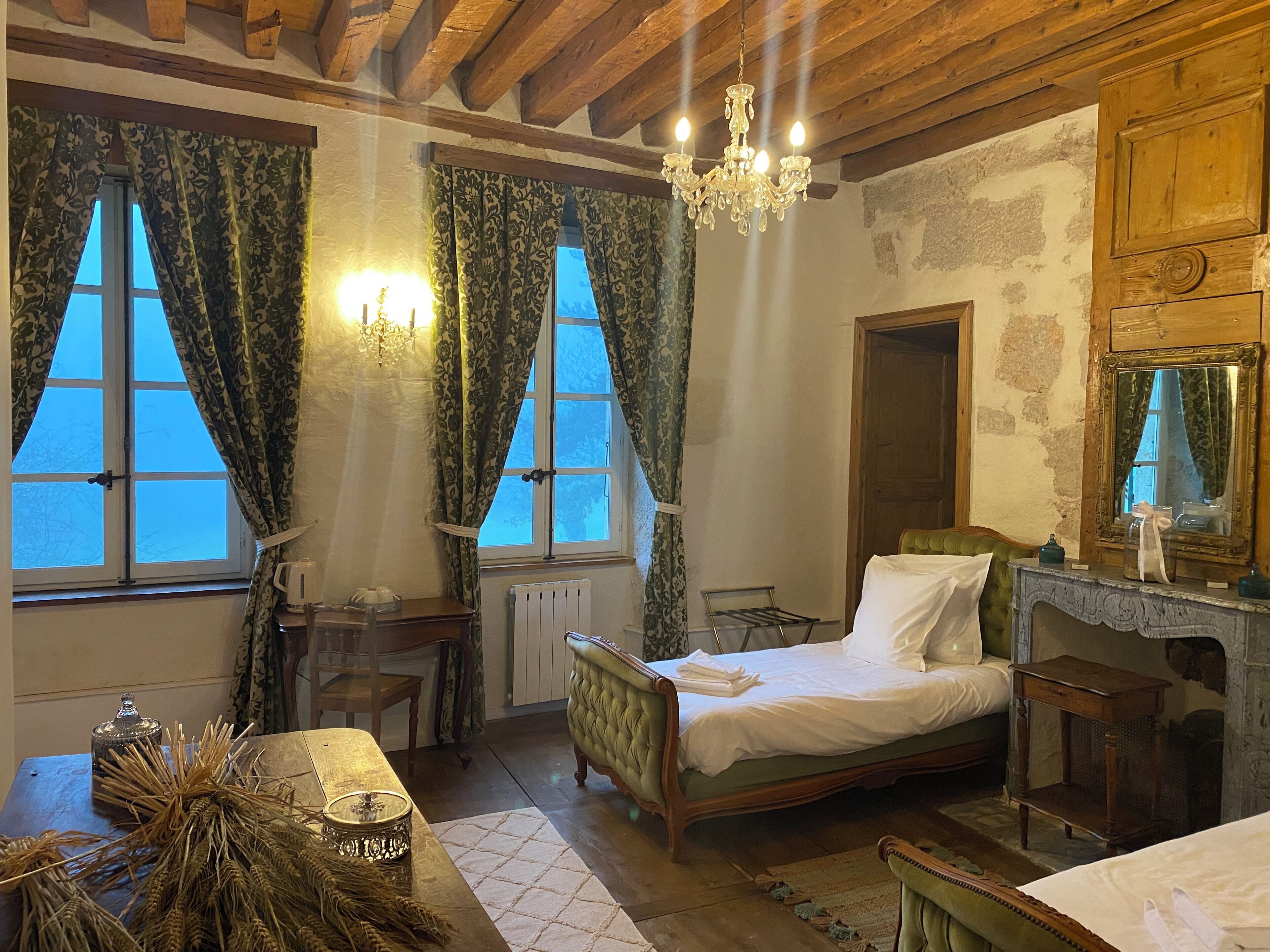 A rustic bedroom with a wooden ceiling, a chandelier, a bed with a green upholstered headboard, a dresser with a mirror, and a window with curtains.