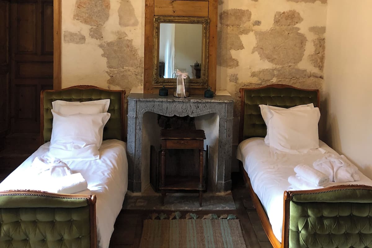 A rustic room with two green upholstered beds, a wooden ceiling, a stone wall, and a chandelier.
