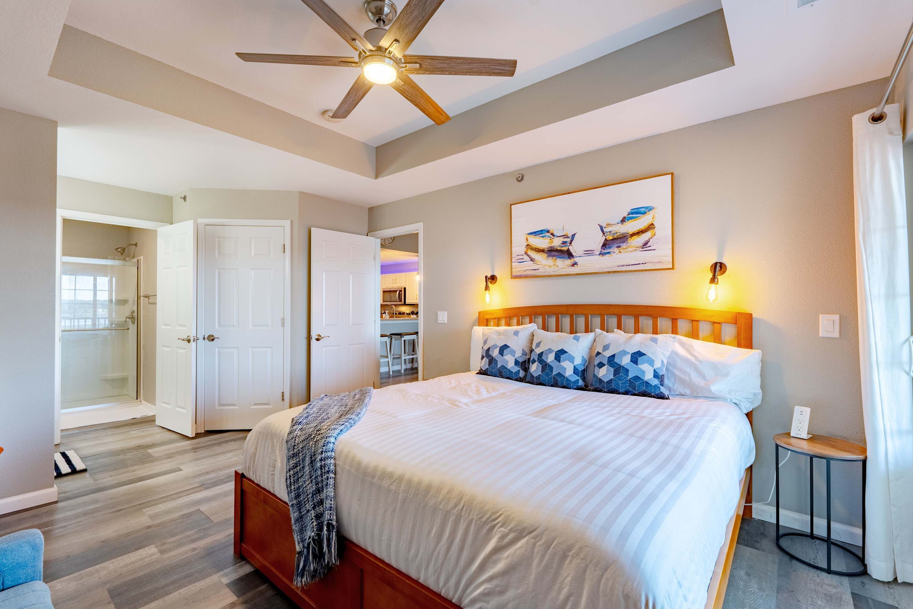A bedroom with a large bed, a ceiling fan, a painting, and a small table.