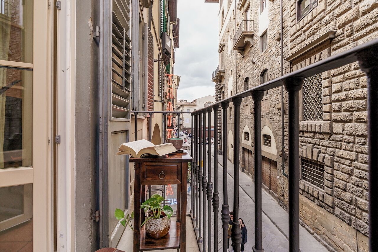 A narrow alleyway with stone buildings, a wooden table with a plant and an open book, and a metal railing.